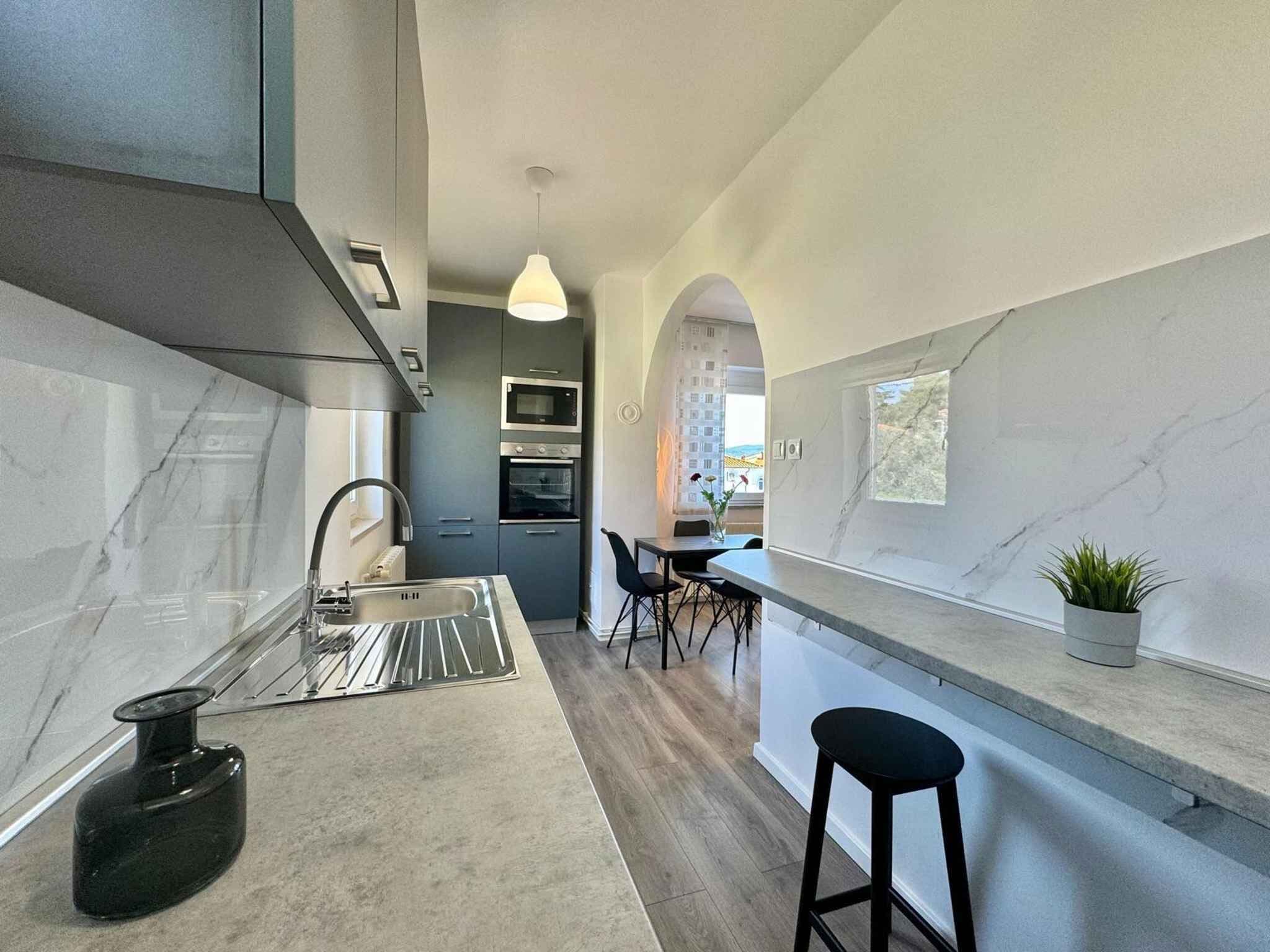 A modern kitchen with a marble countertop, stainless steel appliances, and a view to the outside.