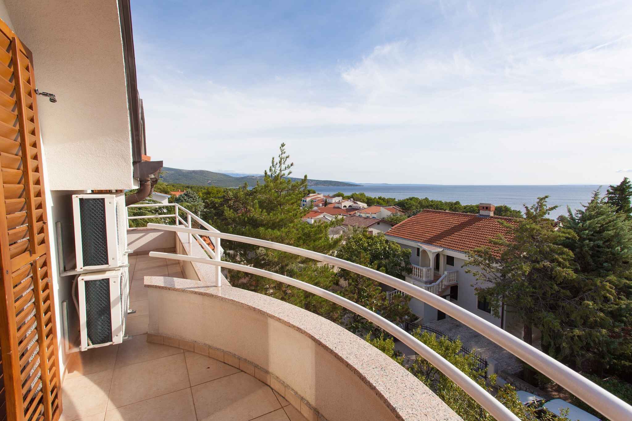 A balcony with a curved railing overlooking a coastal town, a wooden shutter, and an air conditioning unit.