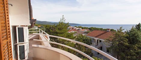 A balcony with a curved railing overlooking a coastal town, a wooden shutter, and an air conditioning unit.