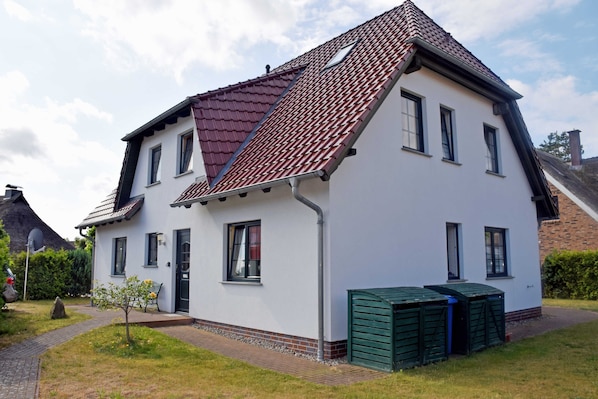 Une maison à deux étages, avec un toit recouvert de tuiles rouges, des murs extérieurs blancs et un petit jardin agrémenté d’un chemin menant à l’entrée.