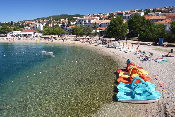Una playa con aguas claras y poco profundas, una fila de botes de alquiler de colores y un pueblo al fondo.