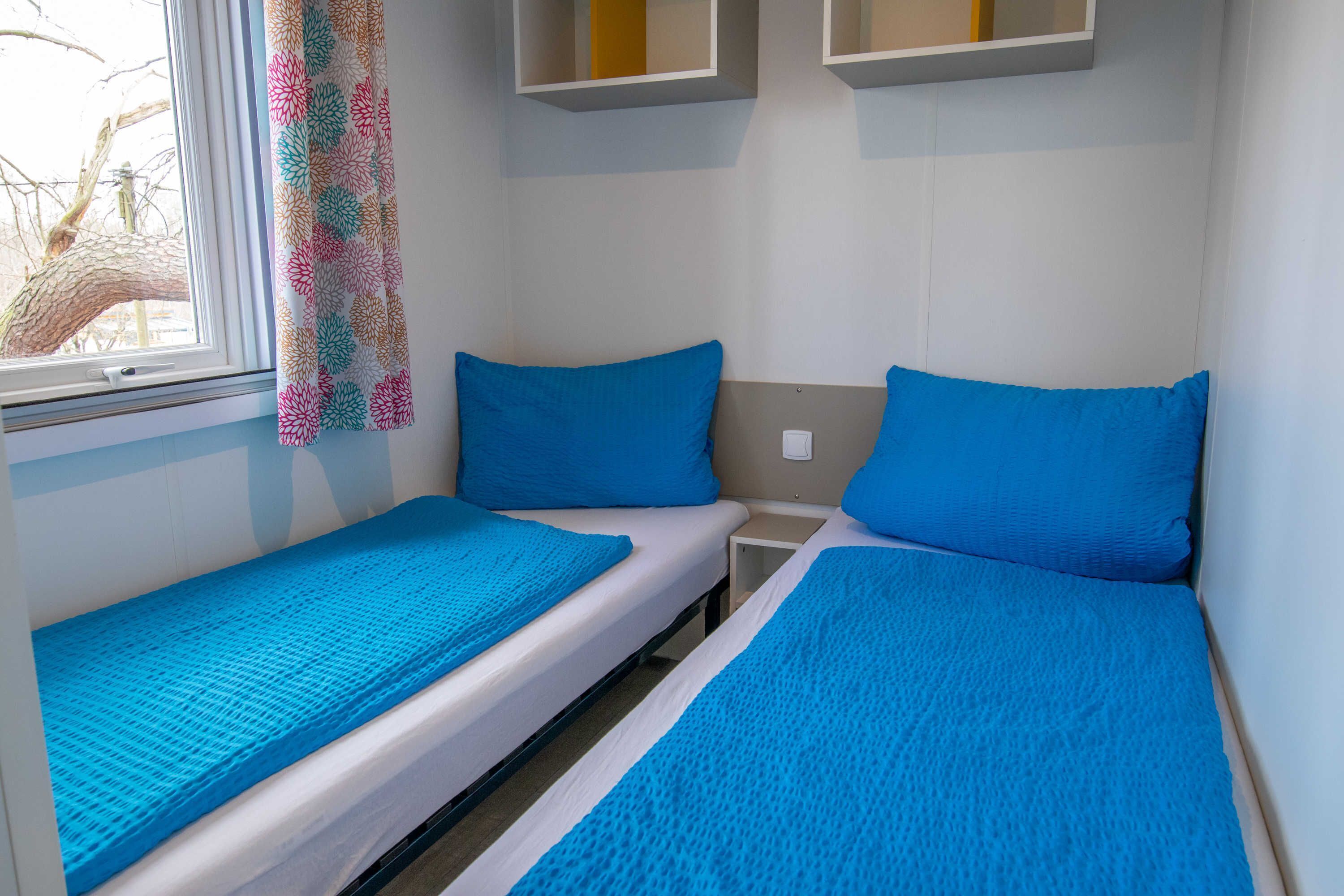A small, compact room with two single beds, each with a blue mattress and pillow. There is a window with a view of bare trees, and a colorful curtain hangs on the window.