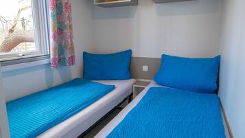 A small, compact room with two single beds, each with a blue mattress and pillow. There is a window with a view of bare trees, and a colorful curtain hangs on the window.