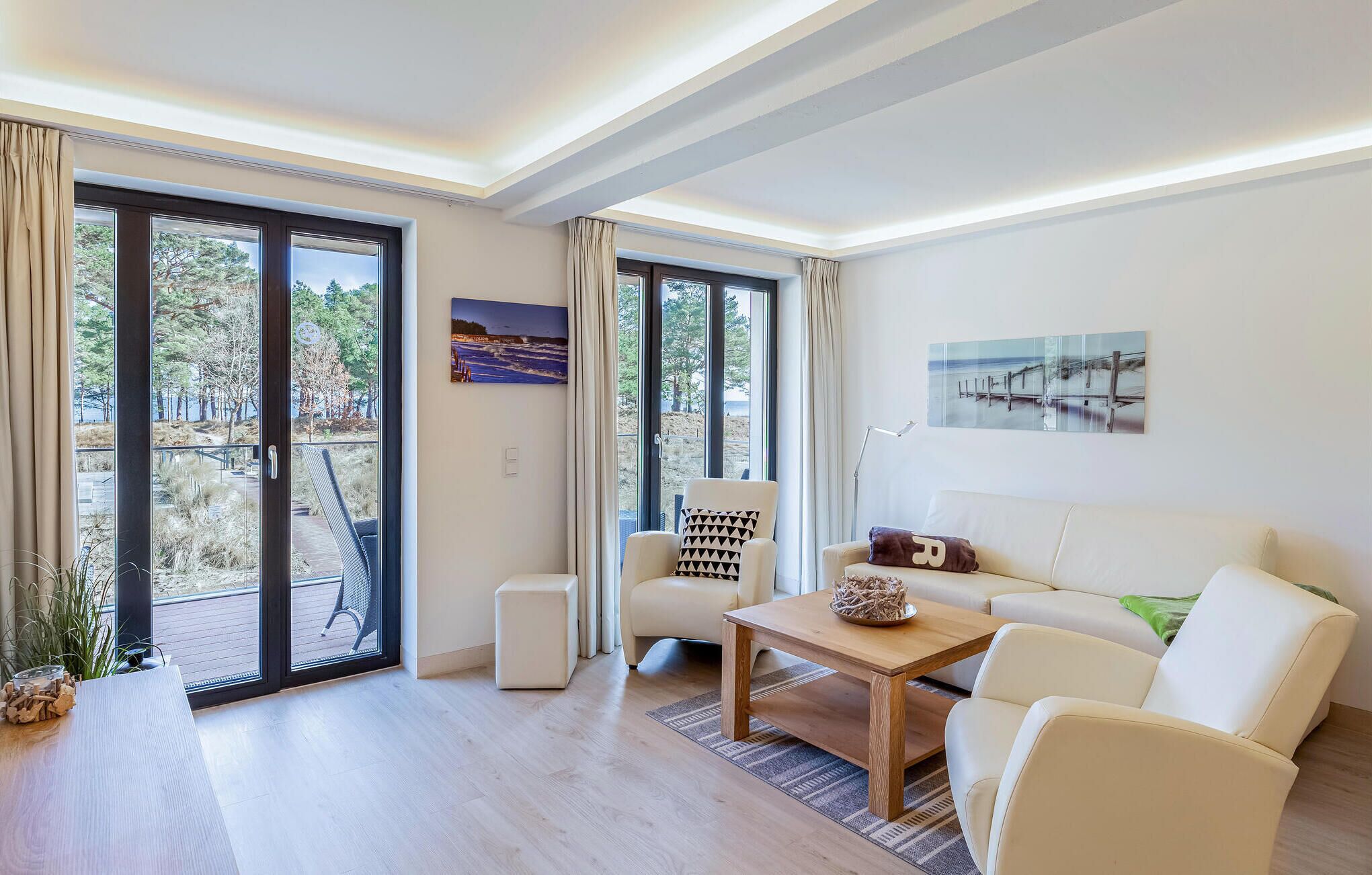 A modern living room with a sofa, armchairs, a coffee table, and large windows offering a view of a beach and trees.
