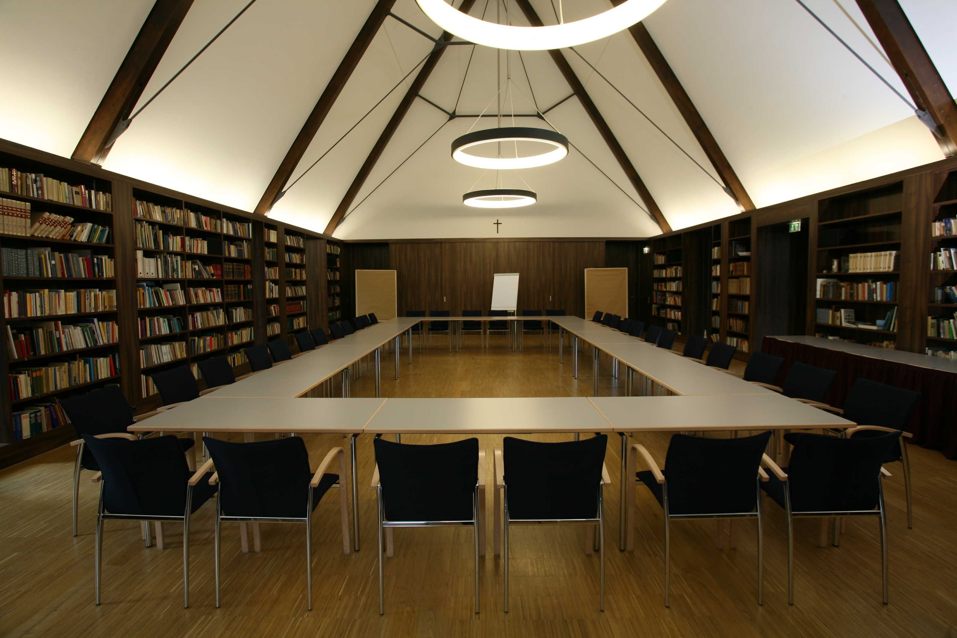 Ett bibliotek med ett långt bord och stolar, trähyllor och ett högt tak med ett kors.