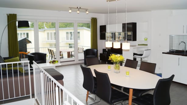 A modern dining area with a round table, black chairs, and a vase of yellow flowers. There is a kitchen area with white cabinets and a sink.