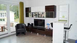 A modern living room with a flat-screen TV, a bookshelf, a dining area with a table and chairs, and a sliding glass door leading to an outdoor balcony.
