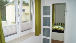 A room with a bed, a window with blinds, a door leading to another room, and a view of a building outside.