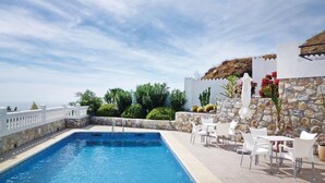 Una piscina con sillas y mesas blancas, rodeada de muros de piedra y plantas.