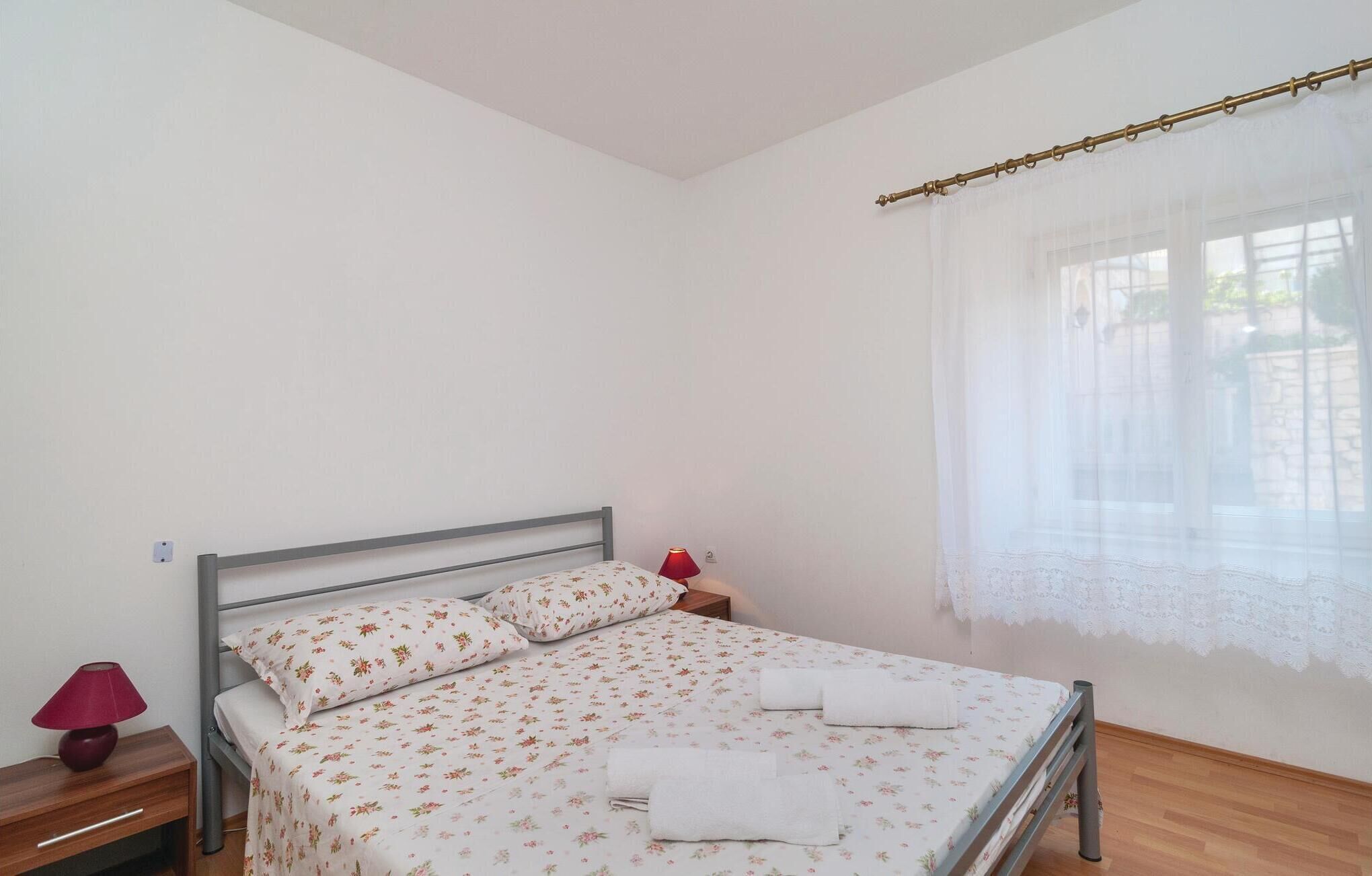 Una cama individual con ropa de cama floral, una lámpara de noche y una ventana con cortinas translúcidas.