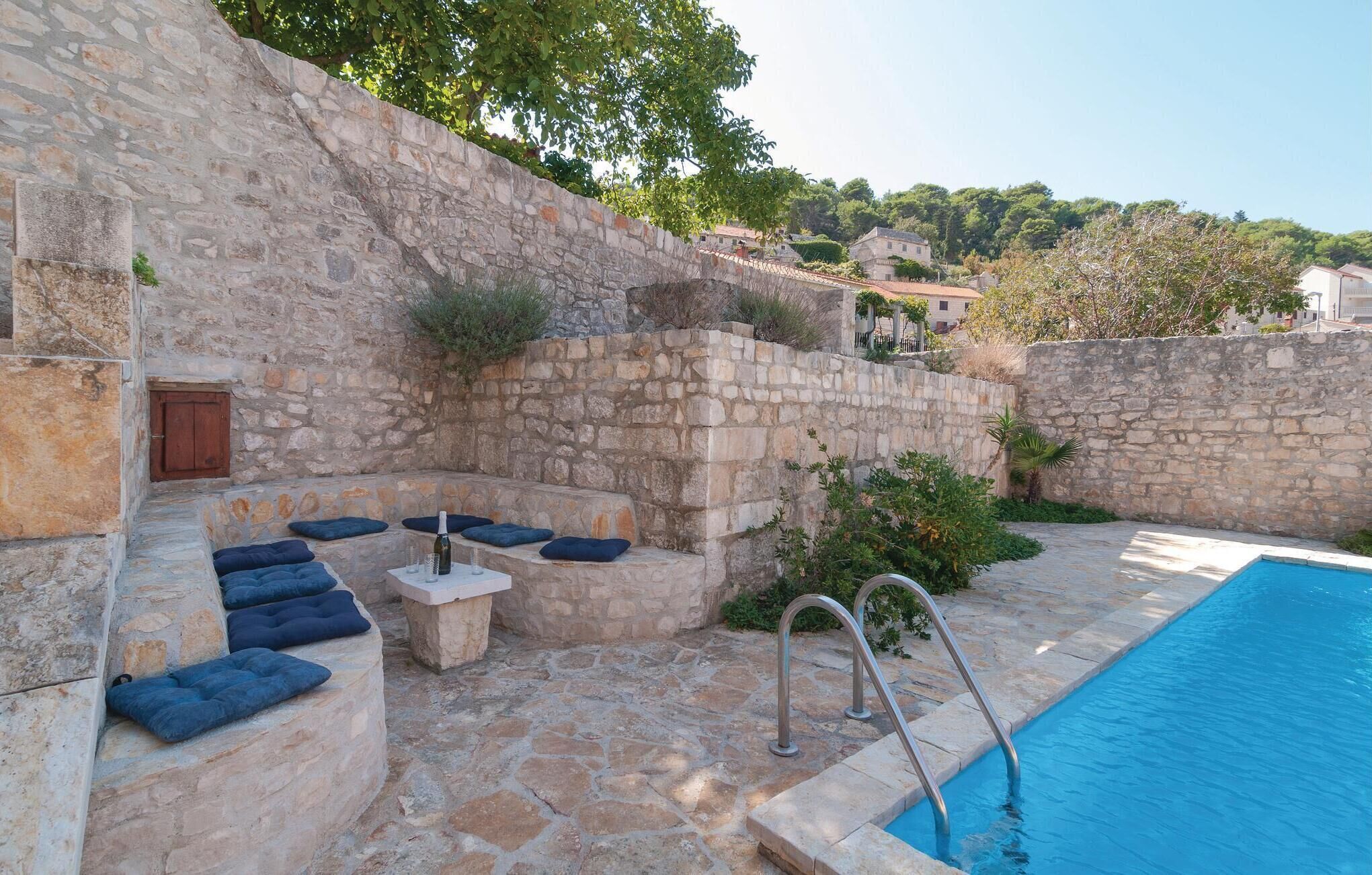 Un espace extérieur aménagé avec des murs en pierre, doté de coussins, d’une petite table et d’une bouteille de vin. On aperçoit une piscine à l’arrière-plan.