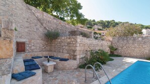 A stone-walled outdoor seating area with cushions, a small table, and a bottle of wine. A pool is visible in the background. - Amazing home in Pucisca (Pucisca)