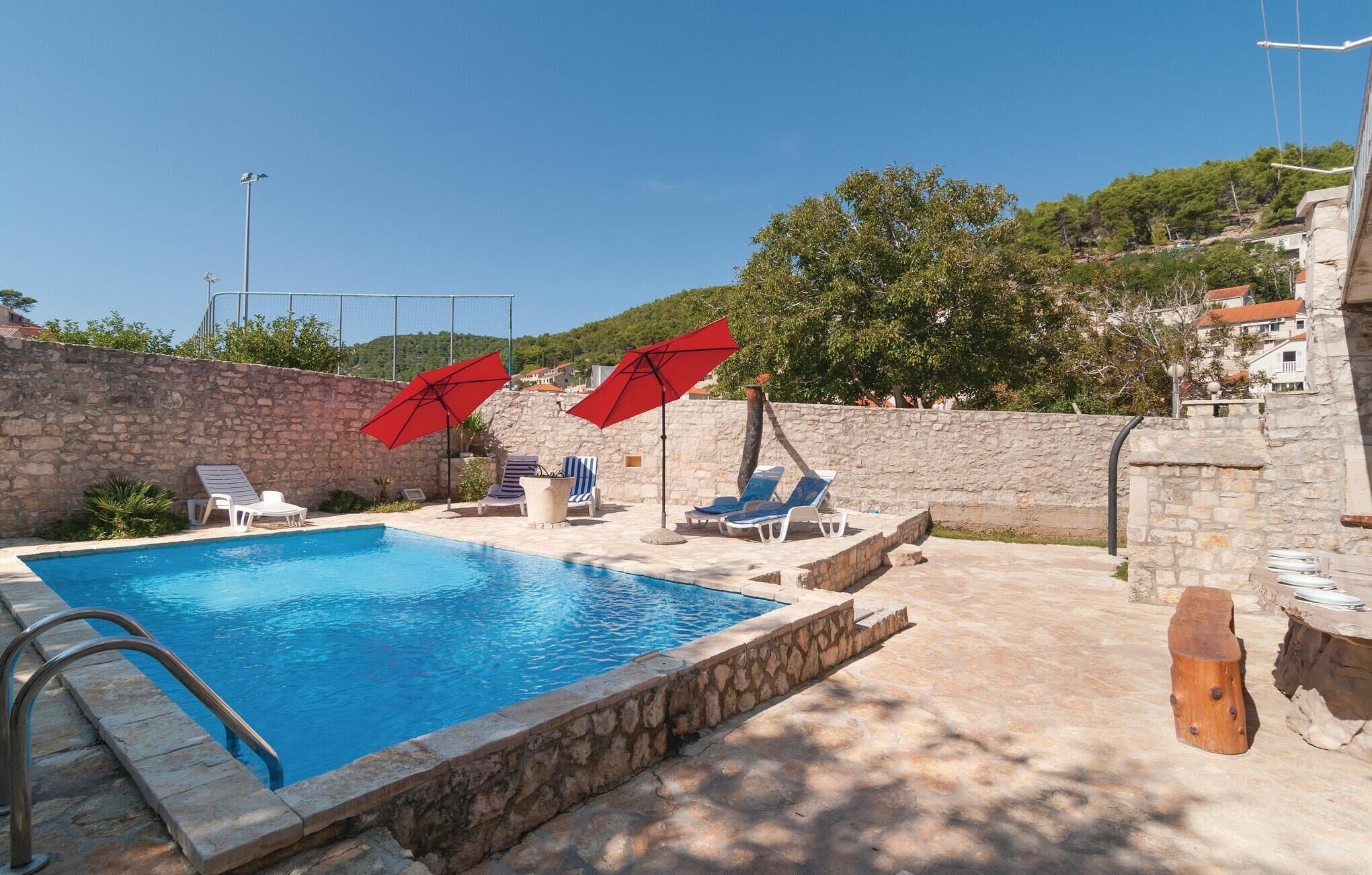 A swimming pool with lounge chairs and red umbrellas, surrounded by stone walls and a view of a hillside.