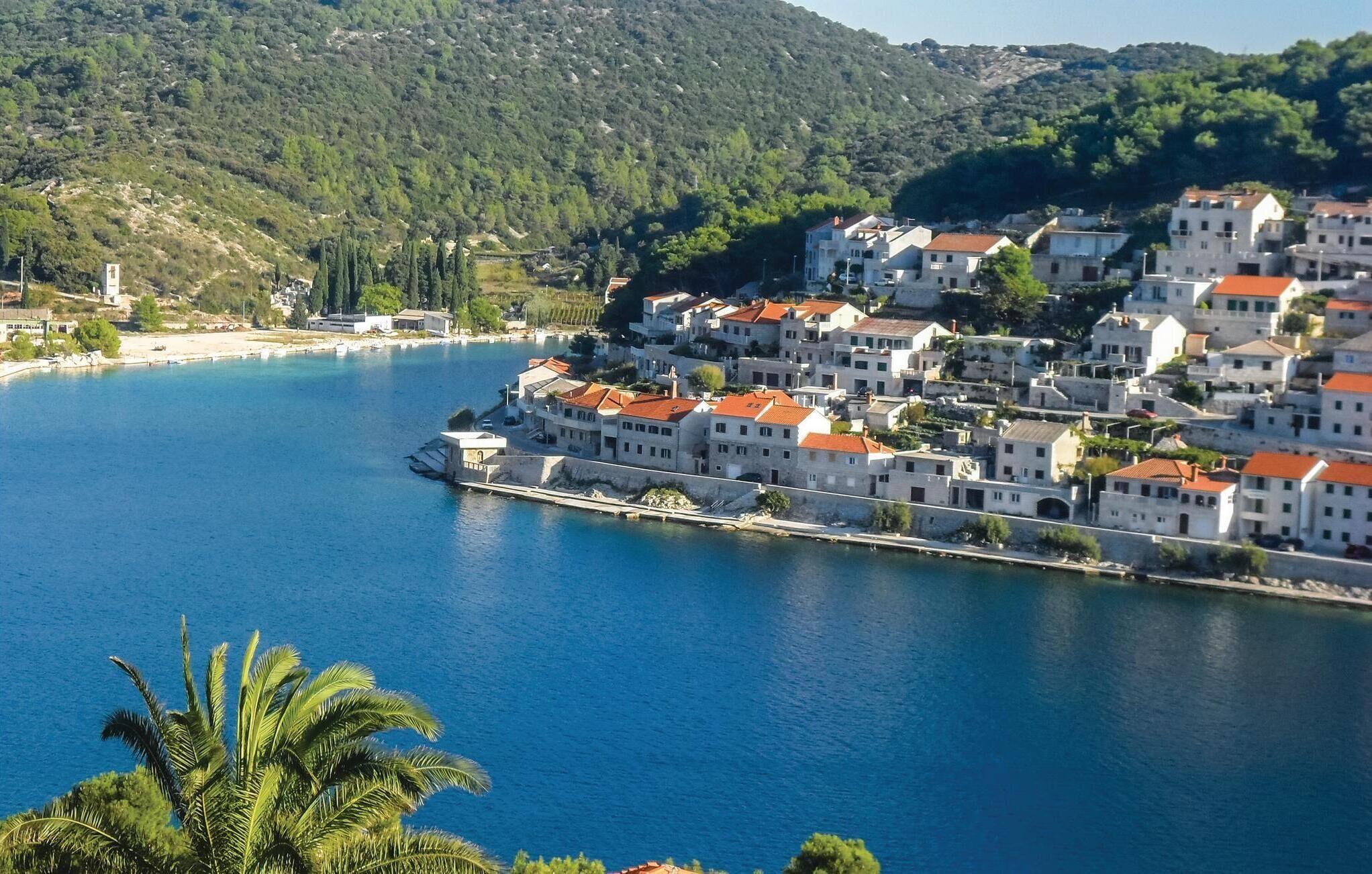 A coastal town with white buildings and red roofs, a clear blue sea, and a hilly backdrop.