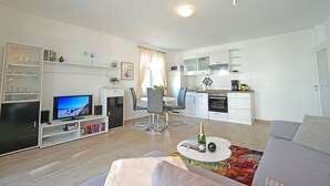 Ein modernes Wohnzimmer mit Flachbildfernseher, einer Couch, einem Couchtisch mit einer Flasche sowie einem Essbereich mit Tisch und Stühlen.