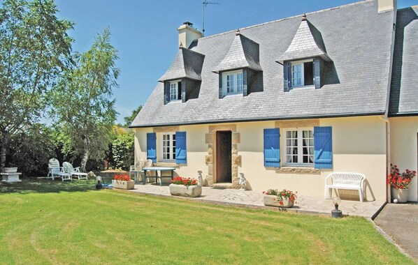 Ein zweistöckiges Haus mit geneigtem Dach, einer Veranda mit weißen Stühlen und einem kleinen Gartenbereich mit Blumentöpfen und Blumen.