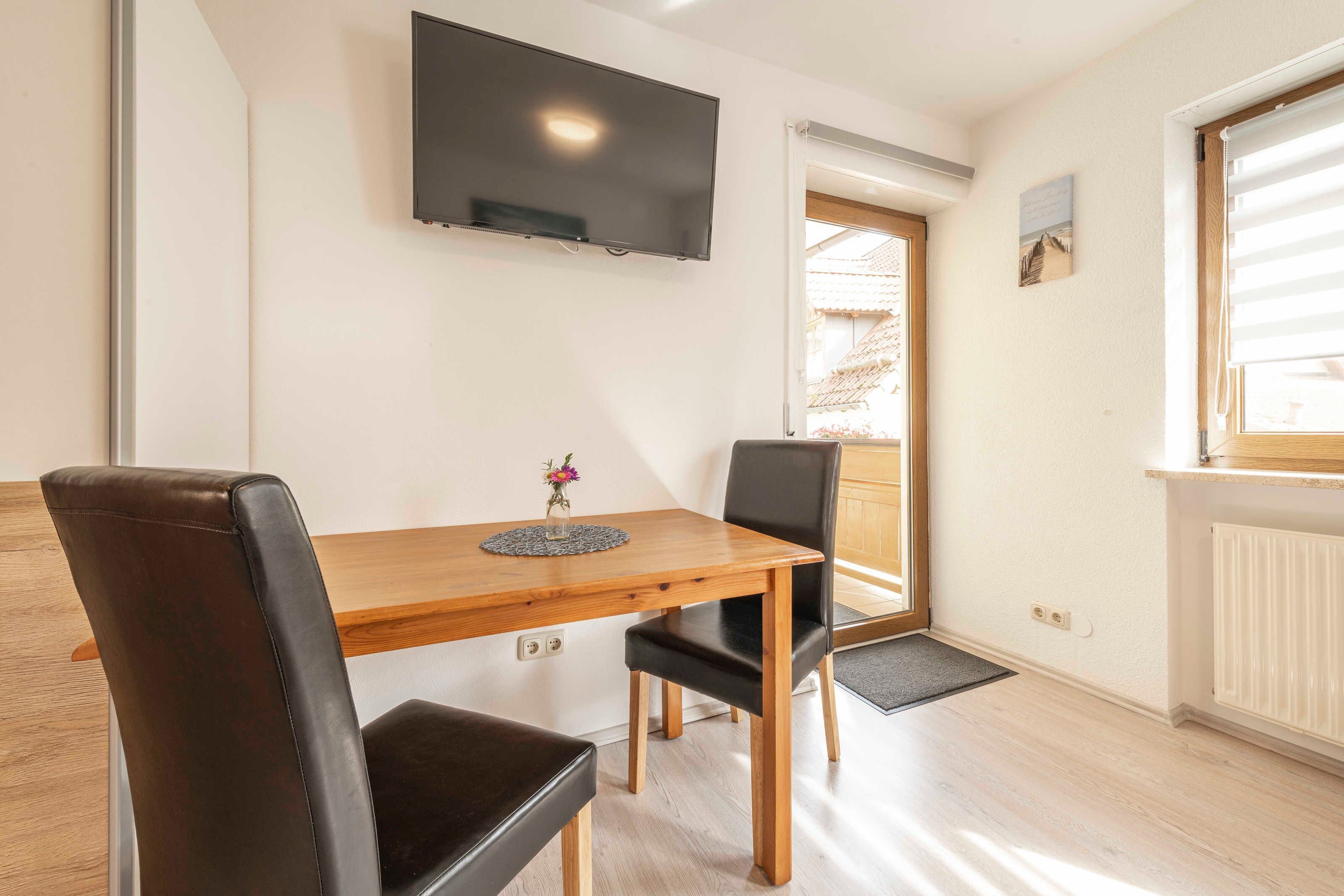 Un comedor con mesa de madera, dos sillas, un florero con flores, televisor montado en la pared y una ventana con persianas.