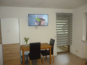 A dining area with a wooden table and chairs, a television mounted on the wall, and a window with blinds. - Double Room - Bed and Breakfast Klein (Heiligenstadt i.OFr.)