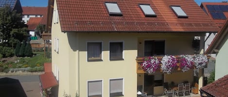 Ein zweistöckiges Haus mit einem geneigten roten Ziegeldach, Solaranlagen und einem Balkon mit hängenden Blumenkörben.