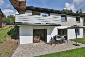 A modern two-story house with a patio area featuring a table and chairs, a large umbrella, and a sliding glass door leading to a balcony.