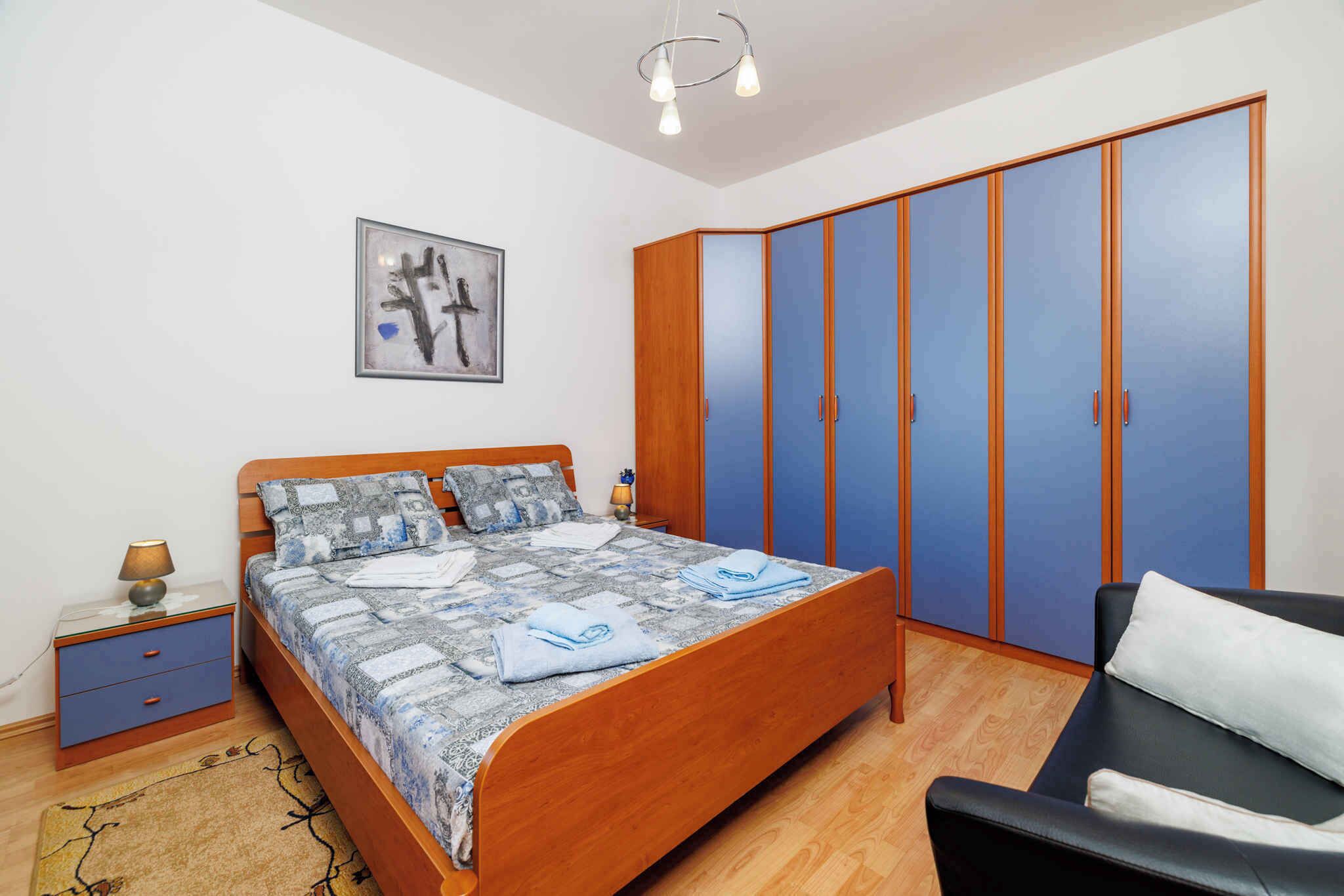 A bedroom with a wooden bed, a blue wardrobe, a black armchair, and a small bedside table with a lamp.