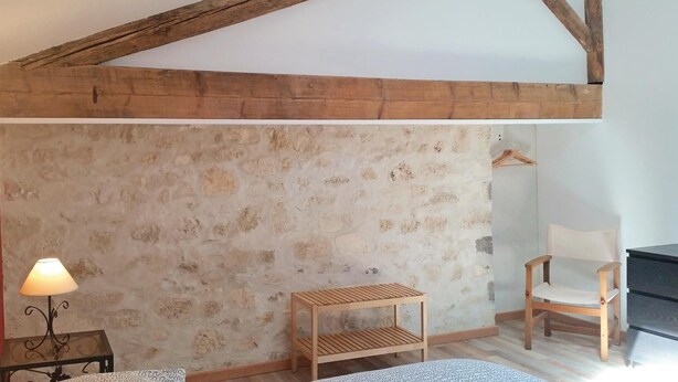 Una habitación rústica con vigas de madera a la vista, un muro de piedra, una silla de madera, una mesita y un perchero colgado.