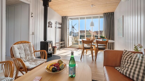 Een knus vertrek met een houten plafond, een zwarte kachel, rieten stoelen, een houten tafel met fruit en een fles, en uitzicht op een molen.