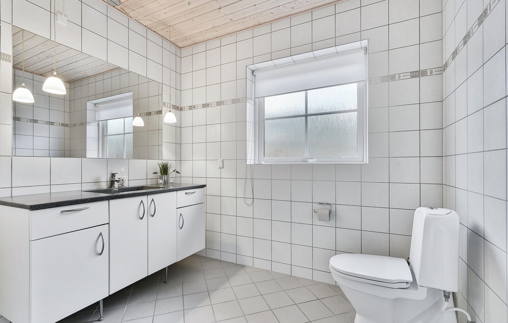 Un baño moderno con azulejos blancos, un lavamanos con espejo, un inodoro e iluminación colgante.
