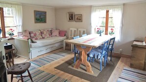 Un salon confortable avec un canapé, une table à manger et des chaises. Il y a deux fenêtres avec des rideaux, un radiateur et un petit tabouret.