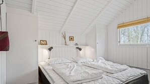 Quarto com cama, duas luminárias de cabeceira, janela com vista para árvores e prateleira com objetos decorativos.