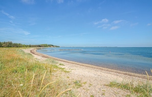 A coastal landscape with a sandy beach, clear blue water, and a distant forested area. - Nice home in Thyholm with kitchen (Thyholm)