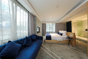 A modern hotel room with a large bed, a sofa, a TV, and a bathroom with a glass partition.