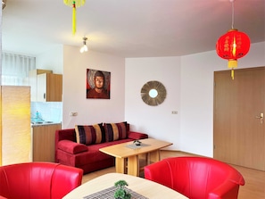 A modern living room with a red sofa, a wooden dining table, and a red chair. There is a kitchen area with a stove and cabinets, and a red lantern hanging from the ceiling.