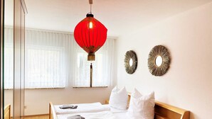Un dormitorio con una cama de madera, ropa de cama blanca y un farol colgante rojo. Hay dos adornos en la pared y una ventana con cortinas de encaje.