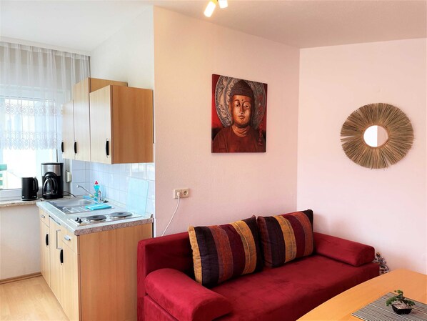 A compact living space with a kitchenette, a red sofa, a wall-mounted Buddha painting, and a decorative mirror.