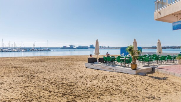 Un café situé en bord de mer, avec une terrasse, une vue sur l’océan et une zone industrielle visible au loin.