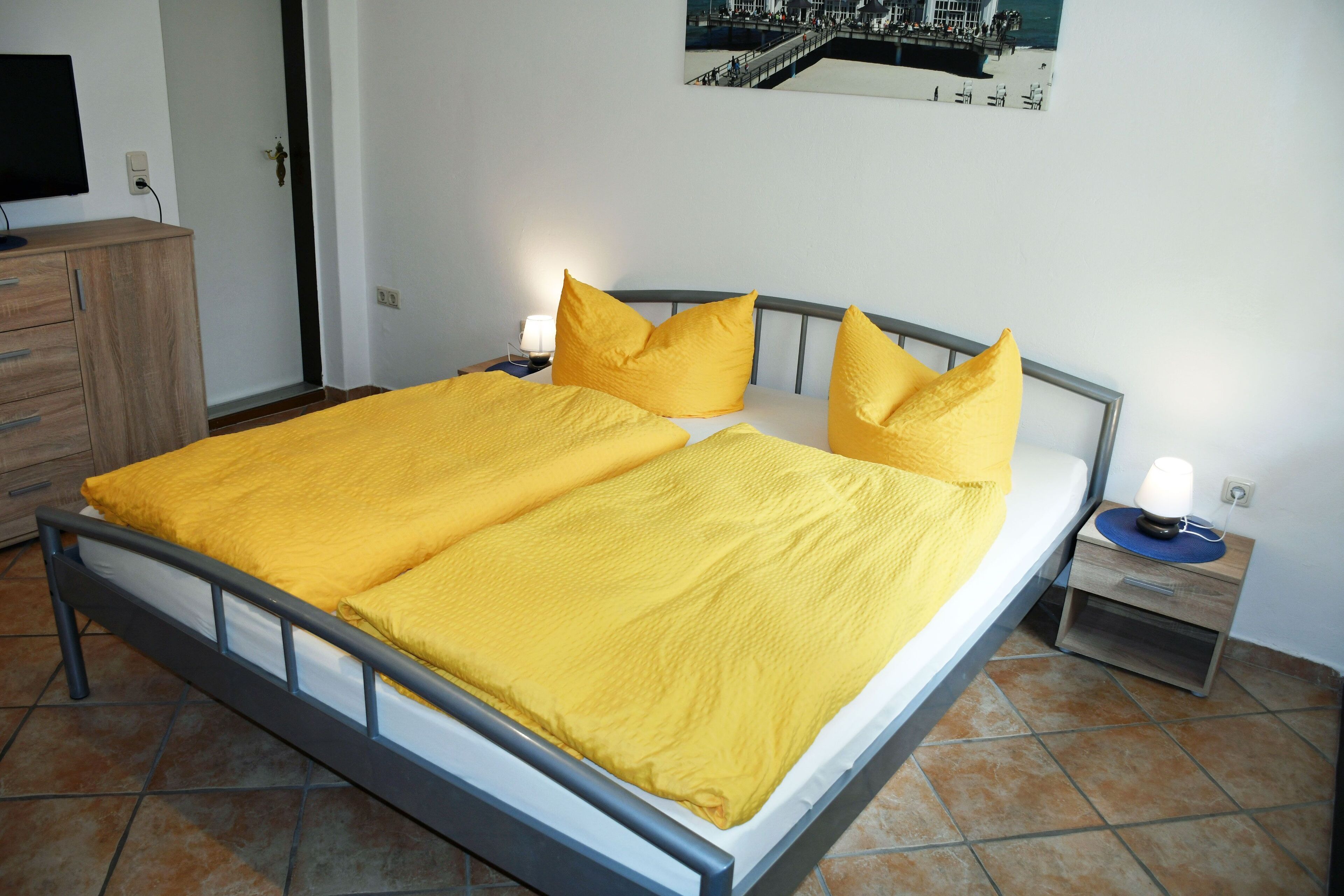 Un letto con struttura in metallo e copripiumino giallo, un comodino con lampada e una TV a parete.