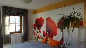 A bedroom with a bed, a radiator, a window with curtains, a wall with a floral mural, and a plant.