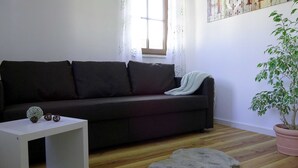 A living room with a dark sofa, a white side table, a potted plant, and a window with floral curtains.