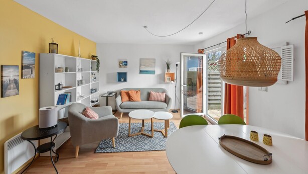 Una sala de estar moderna con un sofá, sillones, una mesa de centro y una zona de comedor con una mesa y sillas. Se observa una estantería, un radiador y una lámpara colgante.