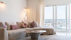 Un salon moderne avec un ensemble de canapés beige, des coussins décoratifs à motifs, une table basse ronde et une vue sur une marina, visible à travers de grandes fenêtres.