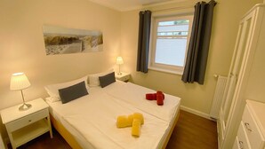 Una habitación de hotel con cama, mesita de noche, lámpara, ventana con cortinas, radiador y un cuadro en la pared.