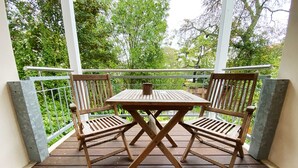 Una terraza de madera con mesa y sillas, rodeada por una barandilla de metal y follaje.
