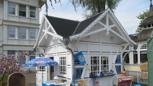 Um quiosque branco com guarda-sol azul, cadeiras de vime e uma mesa com produtos à venda.