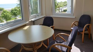 Quarto com uma mesa redonda e quatro cadeiras, grandes janelas com vista para árvores e um corpo d'água, e um radiador sob as janelas.