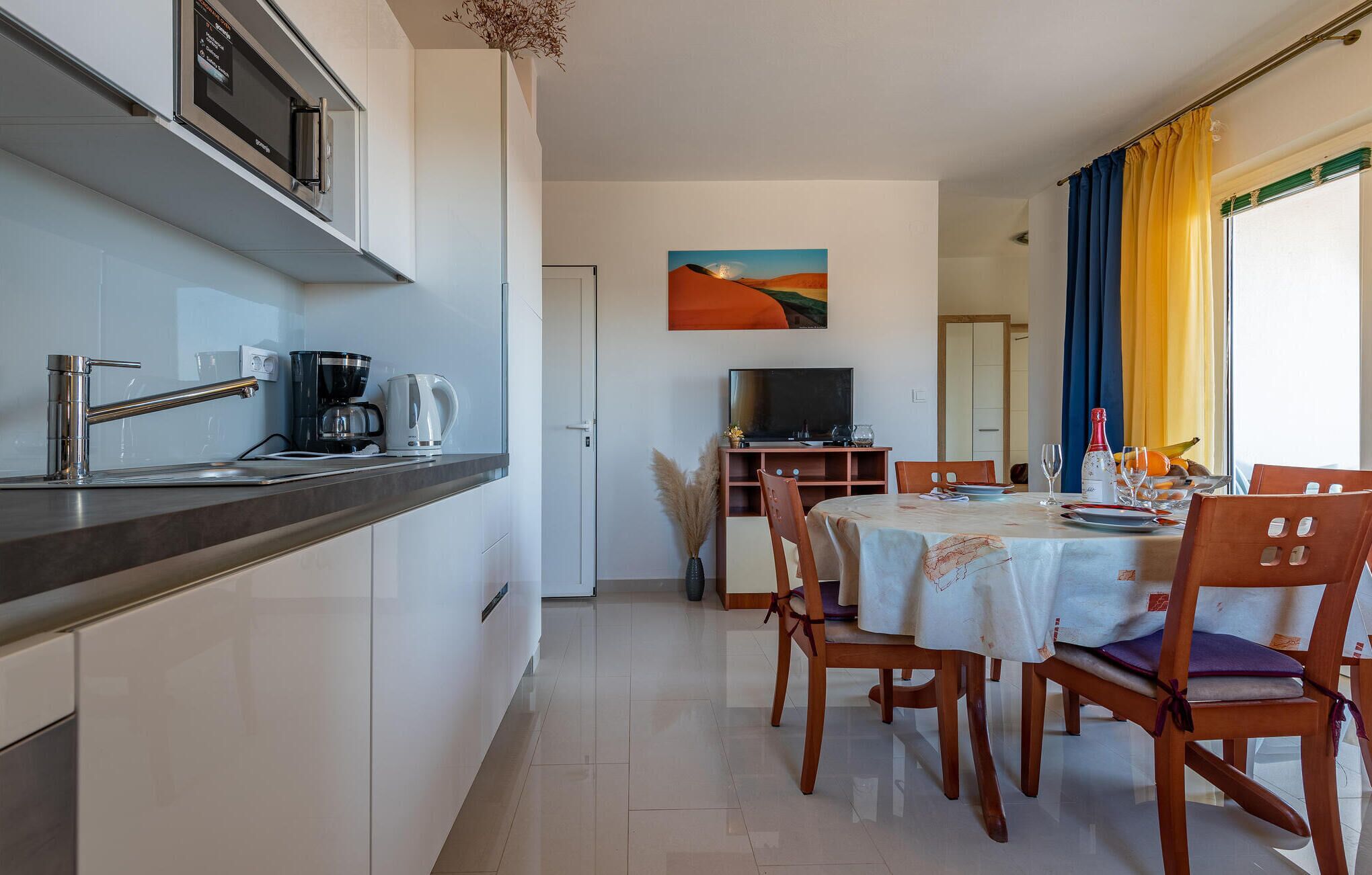 Cocina moderna equipada con microondas, cafetera y hervidor. Hay un comedor con una mesa preparada para dos personas, un televisor y un cuadro en la pared.