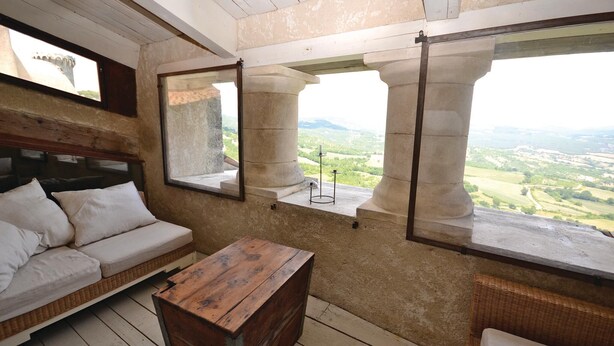 Habitación con un banco de madera, un sofá blanco y vistas a un paisaje a través de amplias ventanas enmarcadas por columnas de piedra.
