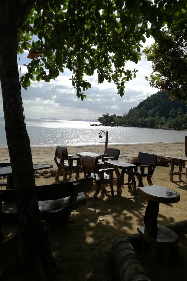 A beachside seating area with tables and chairs, a tree, and a view of the sea and distant hills.