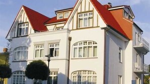 A two-story building with a red roof and white walls, featuring multiple windows and a small tree in front. - (129/3) 2-room apartment - apartment house Plückhahn (Kühlungsborn)