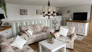 Un salon avec un papier peint à rayures, un lustre, un ensemble de canapés, une table basse et un téléviseur.